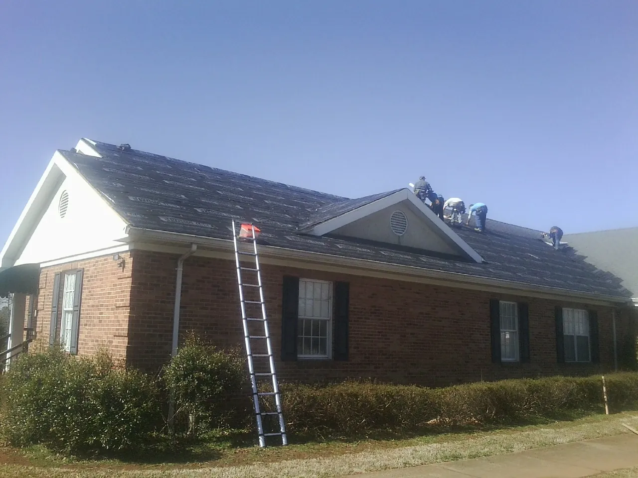 Crew tearing shingles off a brick ranch roof with a ladder set — Metal Roof Repair work by our crew in Walkerton